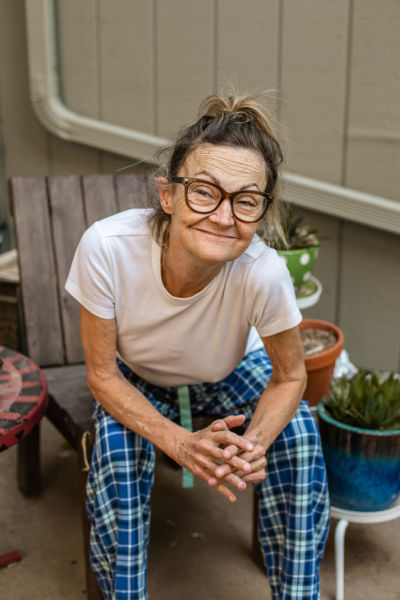 A woman wearing glasses sits with her hands clasped on her front porch