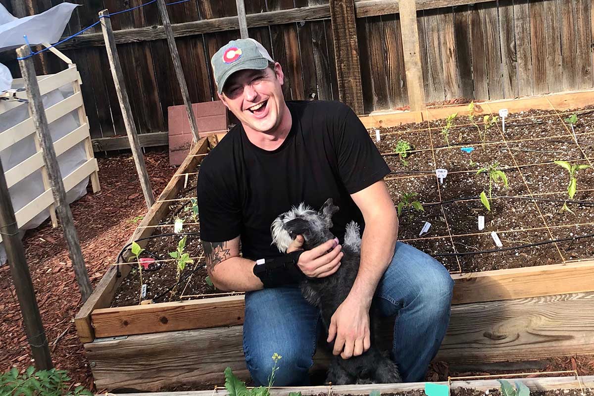 A man sits in his garden with plants sprouting up, petting his dog