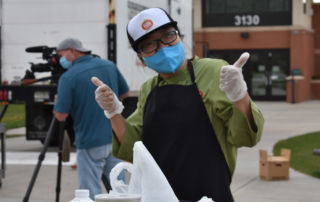 A food worker in a baseball cap and mask gives two thumbs up at a grocery distribution in Boulder
